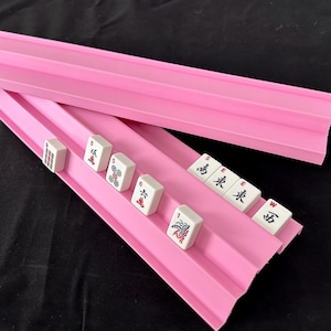 May include: Two bright pink plastic Mahjong tile racks with tiered ledges. One rack is empty, while the other holds several white Mahjong tiles featuring red, green, and black characters, numbers, and symbols, including the letters S, E, W.