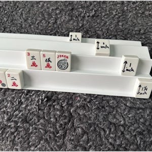 K&ouml;nnte beinhalten: Ein wei&szlig;er Mahjong-Fliesenst&auml;nder aus Kunststoff mit drei Ebenen, der mehrere Mahjong-Fliesen mit roten und schwarzen Schriftzeichen sowie eine Joker-Fliese enth&auml;lt. Einige Fliesen haben handschriftliche Ma&szlig;e von 2,54 cm und 3,18 cm in schwarzer Tinte.