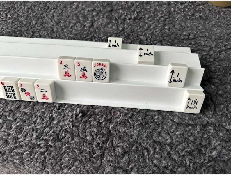 K&ouml;nnte beinhalten: Ein wei&szlig;er Mahjong-Fliesenst&auml;nder aus Kunststoff mit drei Ebenen, der mehrere Mahjong-Fliesen mit roten und schwarzen Schriftzeichen sowie eine Joker-Fliese enth&auml;lt. Einige Fliesen haben handschriftliche Ma&szlig;e von 2,54 cm und 3,18 cm in schwarzer Tinte.