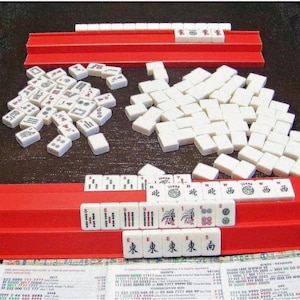 K&ouml;nnte beinhalten: Ein Mahjong-Spiel mit wei&szlig;en Spielsteinen, die schwarze, rote und gr&uuml;ne eingravierte Symbole, Schriftzeichen und Zahlen aufweisen. Das Set enth&auml;lt zwei lange rote Kunststoffhalterungen, einen Haufen verdeckter Steine und eine Spielanleitung.