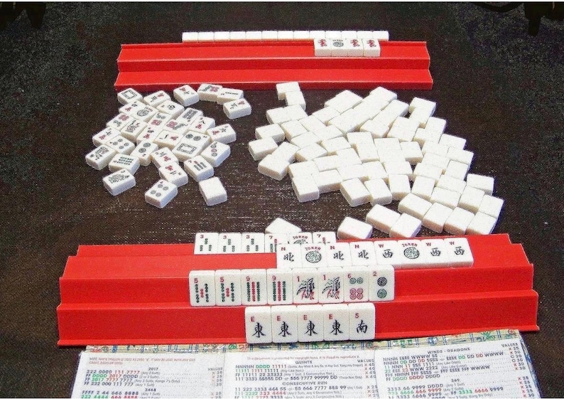 K&ouml;nnte beinhalten: Ein Mahjong-Spiel mit wei&szlig;en Spielsteinen, die schwarze, rote und gr&uuml;ne eingravierte Symbole, Schriftzeichen und Zahlen aufweisen. Das Set enth&auml;lt zwei lange rote Kunststoffhalterungen, einen Haufen verdeckter Steine und eine Spielanleitung.