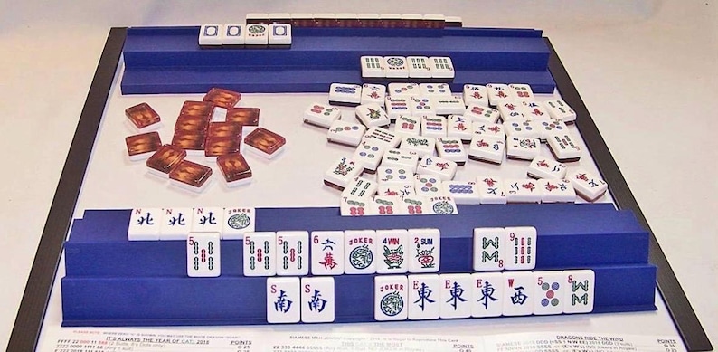 K&ouml;nnte beinhalten: Ein Mahjong-Spielset mit einem wei&szlig;en Spielbrett und blauen, gestuften Ablagen. Das Set enth&auml;lt wei&szlig;e Spielsteine mit verschiedenen schwarzen, roten und gr&uuml;nen Symbolen, Schriftzeichen und Zahlen sowie eine Gruppe brauner Mustersteine.