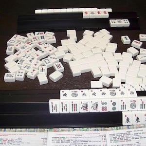 K&ouml;nnte beinhalten: Ein Mahjong-Spiel mit wei&szlig;en rechteckigen Spielsteinen, die schwarze, rote und gr&uuml;ne Symbole, Schriftzeichen und Zahlen tragen. Die Steine liegen auf schwarzen Kunststoffhaltern, erg&auml;nzt durch einen Haufen in der Mitte und eine Punktetafel.