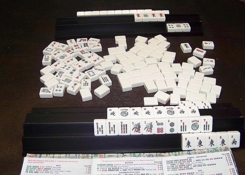 K&ouml;nnte beinhalten: Ein Mahjong-Spiel mit wei&szlig;en rechteckigen Spielsteinen, die schwarze, rote und gr&uuml;ne Symbole, Schriftzeichen und Zahlen tragen. Die Steine liegen auf schwarzen Kunststoffhaltern, erg&auml;nzt durch einen Haufen in der Mitte und eine Punktetafel.