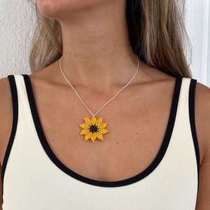 Op de afbeelding: Een wit tanktop met zwarte bies en een zonnebloemketting. De zonnebloem is geel met een bruin centrum.