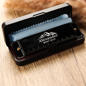 May include: A black harmonica with the text "Alexander BEST MAN" and a mountain graphic, inside a black case with a blue cloth. The harmonica has gold accents and number markings. The case is open, revealing the instrument.