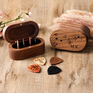 Puede incluir: Una caja de púas de guitarra de madera, abierta para revelar púas, y una caja cerrada con notas musicales y las iniciales "M.K." grabadas en la tapa. Se ven cuatro púas de guitarra, cada una con un diseño y texto únicos.