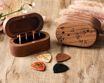 May include: A wooden guitar pick box with a hinged lid, open to reveal picks. The box is next to a closed box with musical notes and the initials "M.K." engraved on the lid. Four guitar picks are visible, each with a unique design and text.