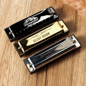 May include: Three harmonicas with personalized text. One is black with "Alexander BEST MAN" and a mountain graphic. Another is gold with "KELSEY HOGAN". The third is silver with "Sherida Dickey RINGBEARER". All have black cases.