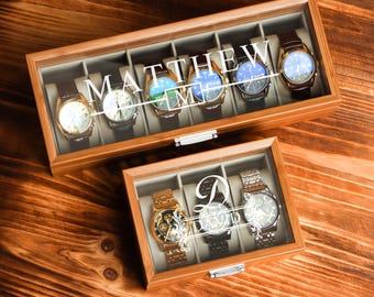 May include: Two wooden watch display cases with glass lids. The top case has the name "MATTHEW" printed on the glass. The cases contain several watches with leather and metal bands. The watches have various face colors and designs. The cases are on a wooden surface.