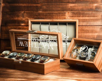 May include: Wooden watch display cases with glass lids. One case is open, displaying six watches with leather bands. The glass lids have engraved names. The cases are on a wooden surface with a wooden background.