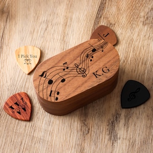 May include: Wooden guitar pick holder with engraved musical notes and initials. Includes several guitar picks with personalized text and a treble clef design. The holder and picks are made of wood and are displayed on a wooden surface.