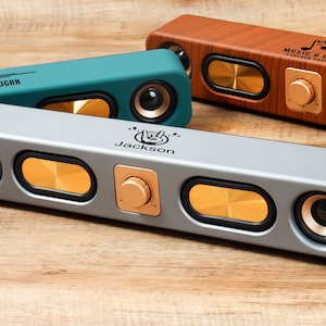 May include: A selection of rectangular speakers in teal, grey, and wood grain finishes. Each speaker has gold-coloured accents, including the speaker cones and control knobs. The grey speaker has the name "Jackson" printed on it.