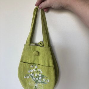 May include: A small, green fabric handbag with a white embroidered lily of the valley design. The bag has a single button closure and two fabric straps.
