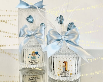 May include: Two glass jars with blue bows and white feathers. The jars have labels with the text "Adam's Baby Shower 08.06.2022" and "Adam's Birthday 08.06.2022".