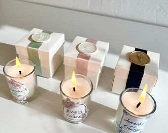 May include: Lit candles in clear glass holders with floral labels and white gift boxes with ribbon and wax seals. The labels read "Jennifer and George" with a date. The boxes have green, pink, and navy blue accents.