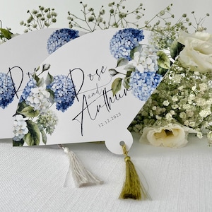 May include: White wedding fan with blue and white hydrangea flowers and green leaves. The fan has a gold tassel and the text "Rose and Anthony 12.12.2025" printed on it.