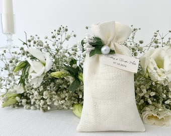May include: A white burlap favor bag with a small bouquet of white flowers and greenery, a pearl accent, and a tag that reads "Brittany & Brian,  Married 07.18.2024".