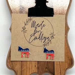 Peut inclure: Une paire de boucles d'oreilles clous en forme d'âne avec des étoiles rouges, blanches et bleues. Les boucles d'oreilles sont présentées sur un presse-papiers en bois avec une carte en papier brun qui dit "Made by Emiliza".