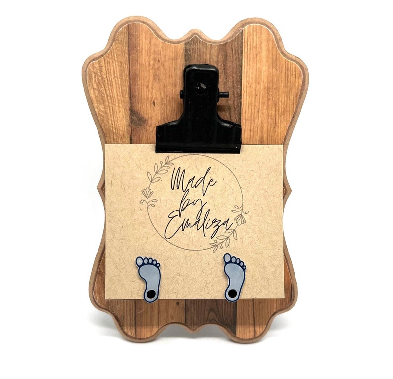 May include: A wooden clipboard with a black clip and a brown card with the text "Made by Emiliza" in a circle with floral accents. Two blue baby footprints are attached to the bottom of the card.