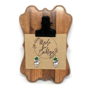 Puede incluir: Portapapeles de madera con un clip negro que sujeta una tarjeta marrón con el texto "Made by Emaliza". Dos pequeños pendientes de alienígenas de colores están adheridos a la tarjeta. El portapapeles tiene un patrón de vetas de madera.