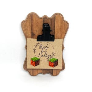 Puede incluir: Portapapeles de madera con un clip negro que sujeta una tarjeta de papel kraft. La tarjeta presenta el texto "Made by Emiliza" y dos pendientes cúbicos pixelados con colores verdes y marrones.
