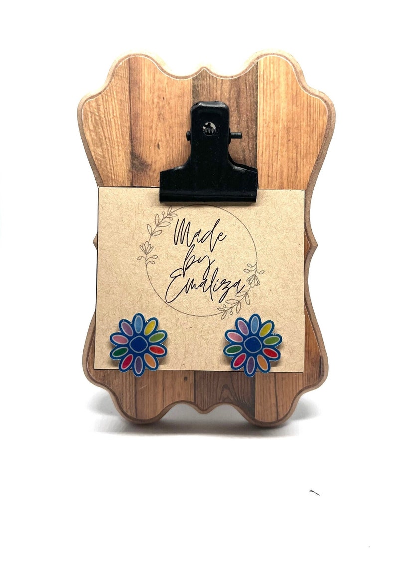 May include: A pair of colorful flower stud earrings on a wooden display stand with a sign that says "Made by Emiliza".