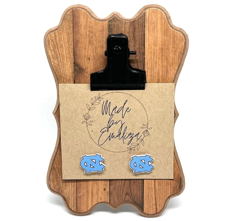 May include: A pair of light blue UNC Tar Heels logo earrings on a brown wooden clipboard with a card that says "Made by Emiliza".