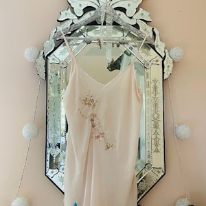 May include: A pale pink, sleeveless slip dress with delicate floral embroidery hanging on a silver ornate mirror with white pom-poms strung around it.