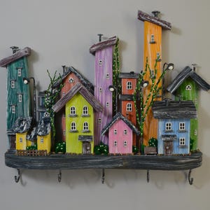 May include: A colorful wooden key holder featuring a whimsical village scene. The houses are painted in various colors, including green, yellow, pink, and blue, with decorative details and hooks for keys. The key holder is mounted on a wooden shelf.
