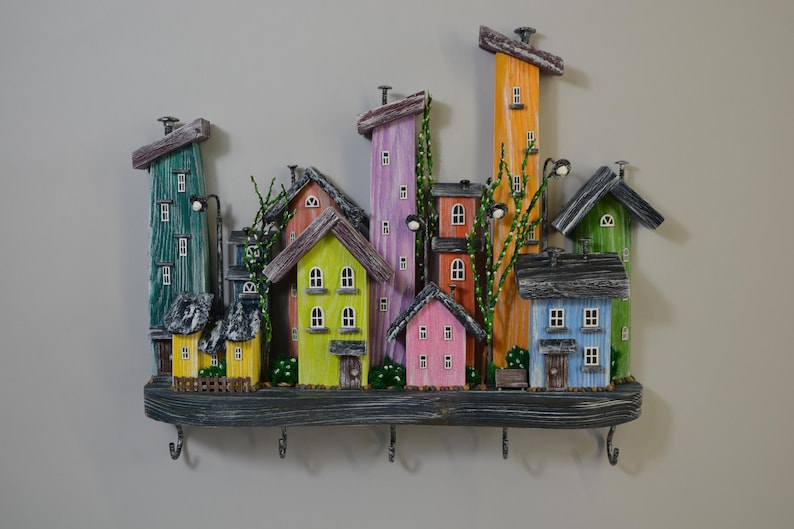 May include: A colorful wooden key holder featuring a whimsical village scene. The houses are painted in various colors, including green, yellow, pink, and blue, with decorative details and hooks for keys. The key holder is mounted on a wooden shelf.