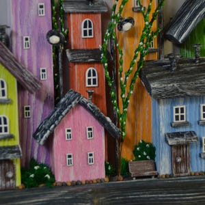 May include: A colorful wooden village scene featuring houses in shades of yellow, pink, orange, purple, blue, and green. Each house has a dark gray roof and white-framed windows. Decorative street lamps and greenery add detail to the whimsical artwork.