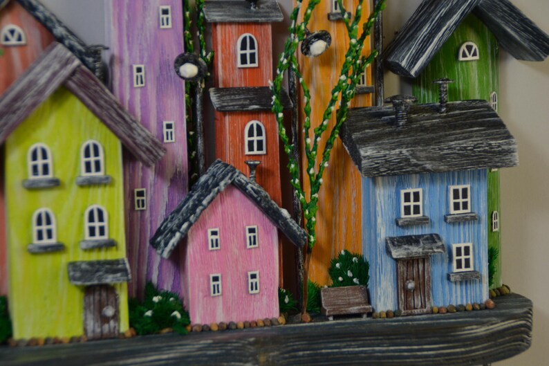 May include: A colorful wooden village scene featuring houses in shades of yellow, pink, orange, purple, blue, and green. Each house has a dark gray roof and white-framed windows. Decorative street lamps and greenery add detail to the whimsical artwork.