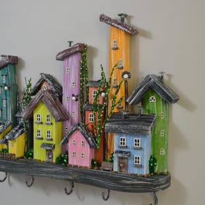 May include: A colorful, whimsical wooden key holder shaped like a village. The houses are painted in various colors, including yellow, pink, green, and blue, with dark gray roofs. The key holder has several metal hooks and is mounted on a gray wooden shelf.