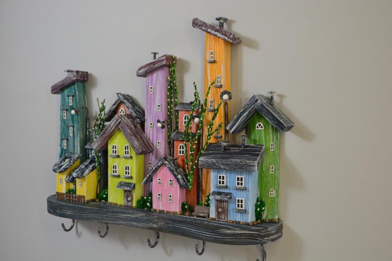 May include: A colorful, whimsical wooden key holder shaped like a village. The houses are painted in various colors, including yellow, pink, green, and blue, with dark gray roofs. The key holder has several metal hooks and is mounted on a gray wooden shelf.