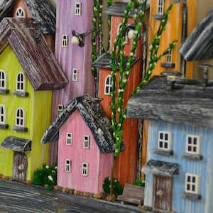 May include: A colorful wooden village scene featuring miniature houses in various hues, including yellow, pink, blue, and orange. Each house has a textured roof, windows, and a door. Green foliage and small benches add detail to the whimsical artwork.