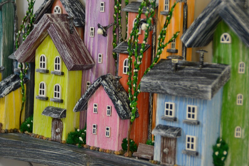 May include: A colorful wooden village scene featuring miniature houses in various hues, including yellow, pink, blue, and orange. Each house has a textured roof, windows, and a door. Green foliage and small benches add detail to the whimsical artwork.
