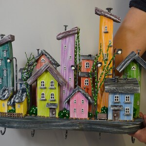 May include: A colorful wooden wall-mounted key holder featuring a whimsical village scene. The houses are painted in various colors, including green, yellow, pink, and orange, with small windows and decorative details. The key holder has several hooks.