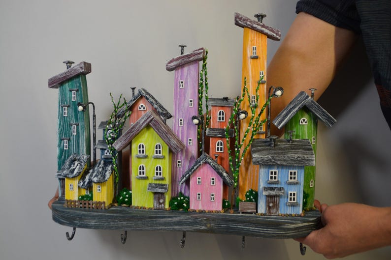 May include: A colorful wooden wall-mounted key holder featuring a whimsical village scene. The houses are painted in various colors, including green, yellow, pink, and orange, with small windows and decorative details. The key holder has several hooks.
