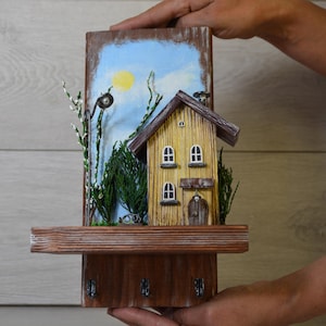 Può includere: Un portachiavi in legno con una casa in miniatura con facciata gialla, tetto marrone e piccole finestre. Lo sfondo raffigura un cielo blu con un sole giallo. Ha un ripiano e ganci per chiavi.