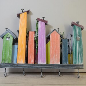 May include: A colorful wooden key holder shaped like a cityscape. The design features various painted buildings in shades of green, orange, blue, and purple. It includes hooks for keys and decorative elements.