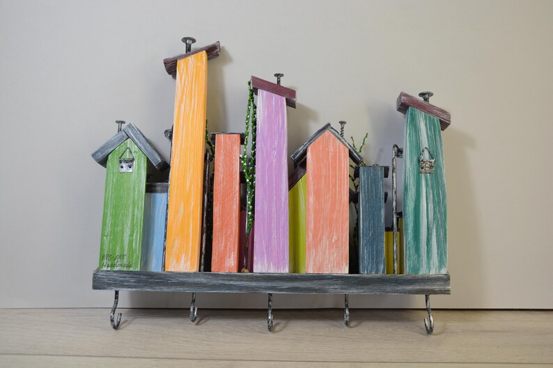 May include: A colorful wooden key holder shaped like a cityscape. The design features various painted buildings in shades of green, orange, blue, and purple. It includes hooks for keys and decorative elements.