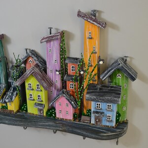 May include: A colorful wooden wall-mounted key holder featuring a whimsical cityscape design. The houses are painted in various bright colors, including yellow, pink, green, and orange, with gray roofs. The key holder has several hooks for keys.