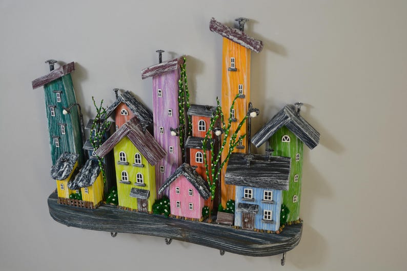 May include: A colorful wooden wall-mounted key holder featuring a whimsical cityscape design. The houses are painted in various bright colors, including yellow, pink, green, and orange, with gray roofs. The key holder has several hooks for keys.