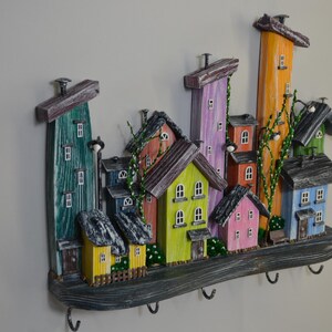 May include: A colorful, wooden wall-mounted key holder featuring a whimsical village scene. The houses are painted in various colors, including green, yellow, pink, and orange, with dark gray roofs. The key holder has five metal hooks.