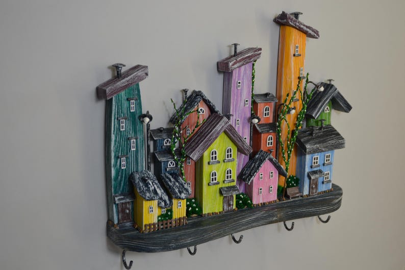 May include: A colorful, wooden wall-mounted key holder featuring a whimsical village scene. The houses are painted in various colors, including green, yellow, pink, and orange, with dark gray roofs. The key holder has five metal hooks.