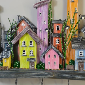 May include: A colorful wooden wall-mounted key holder featuring a whimsical village scene. The houses are painted in various bright colors, including green, yellow, pink, and blue, with small windows and decorative details. The key holder has several hooks.