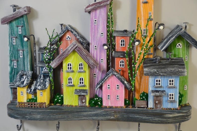 May include: A colorful wooden wall-mounted key holder featuring a whimsical village scene. The houses are painted in various bright colors, including green, yellow, pink, and blue, with small windows and decorative details. The key holder has several hooks.