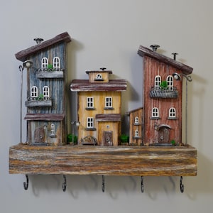 Könnte beinhalten: Ein dekorativer Schlüsselhalter aus Holz mit einer Miniatur-Dorfszene. Die Häuser sind in Blau-, Gelb- und Rottönen bemalt und haben kleine Fenster und Balkone. Der Schlüsselhalter hat fünf Metallhaken und ist auf einer Holzplanke montiert.