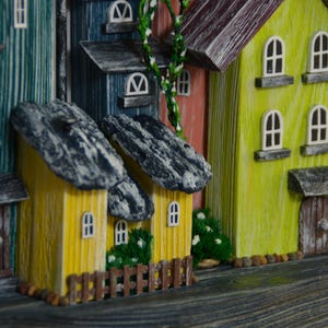 May include: A colorful, handcrafted miniature village scene. The houses are painted in various hues, including yellow, blue, and pink, with stone-like roofs. Small windows and doors add detail, and miniature greenery is placed around the houses.